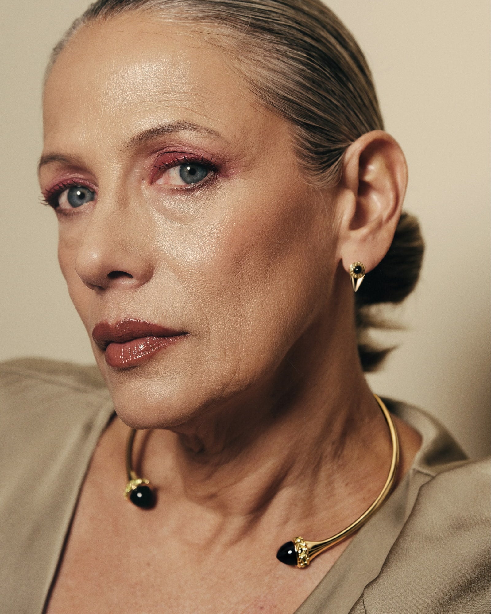 A close-up portrait of a woman wearing a gold-toned c-shaped collar necklace with black bead accents and matching stud earrings, against a warm beige background.