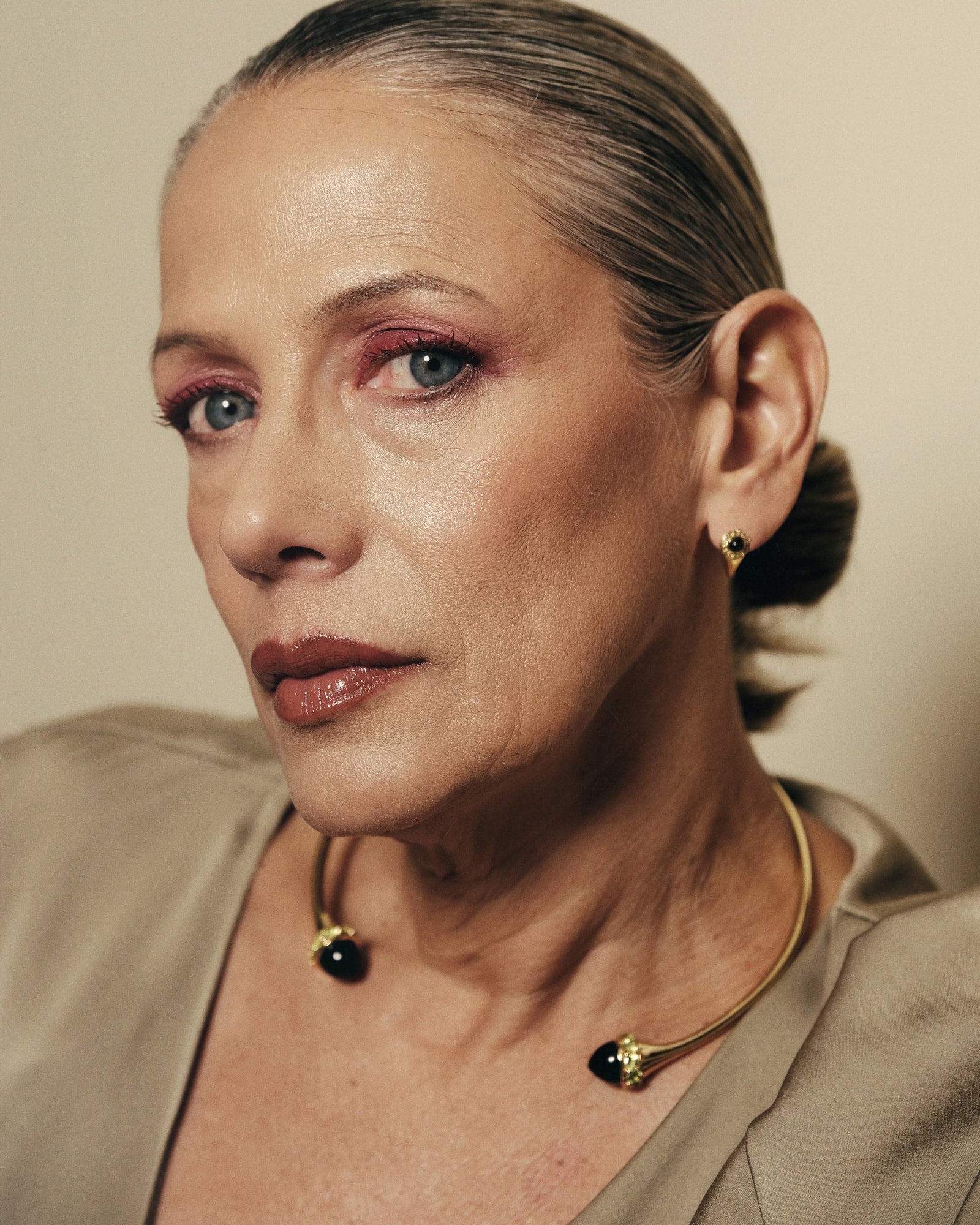 A close-up portrait of a woman with slicked-back hair wearing a beige satin top and gold jewelry with black bead accents against a warm beige background.