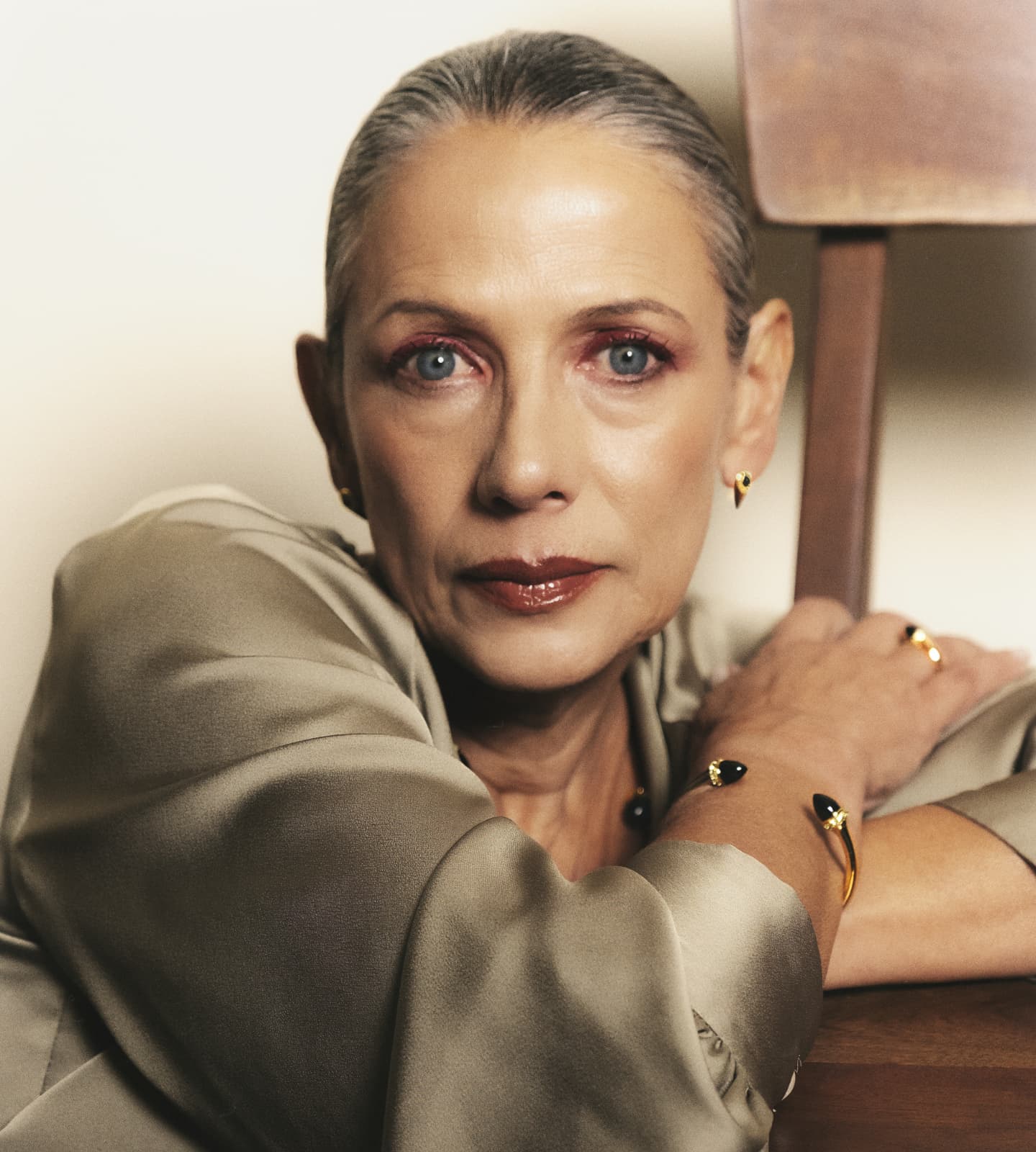 Woman wearing a beige outfit with jewelry, sitting indoors.