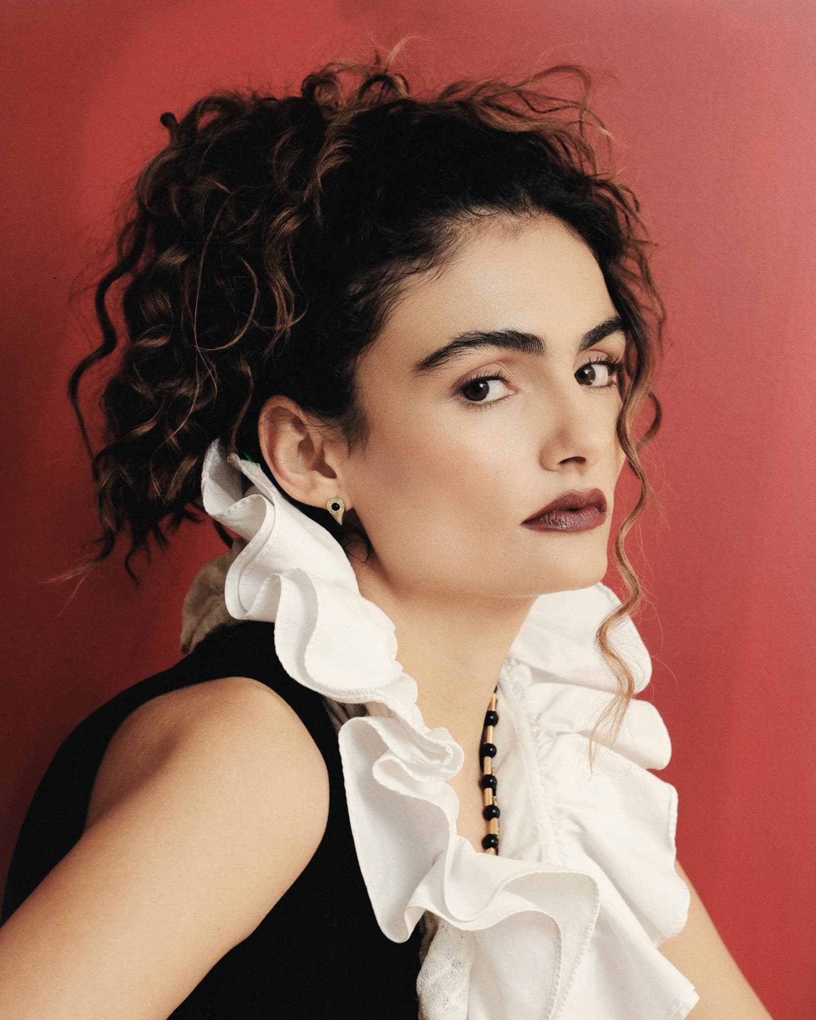 Woman with styled hair wearing a white ruffled blouse against a red background