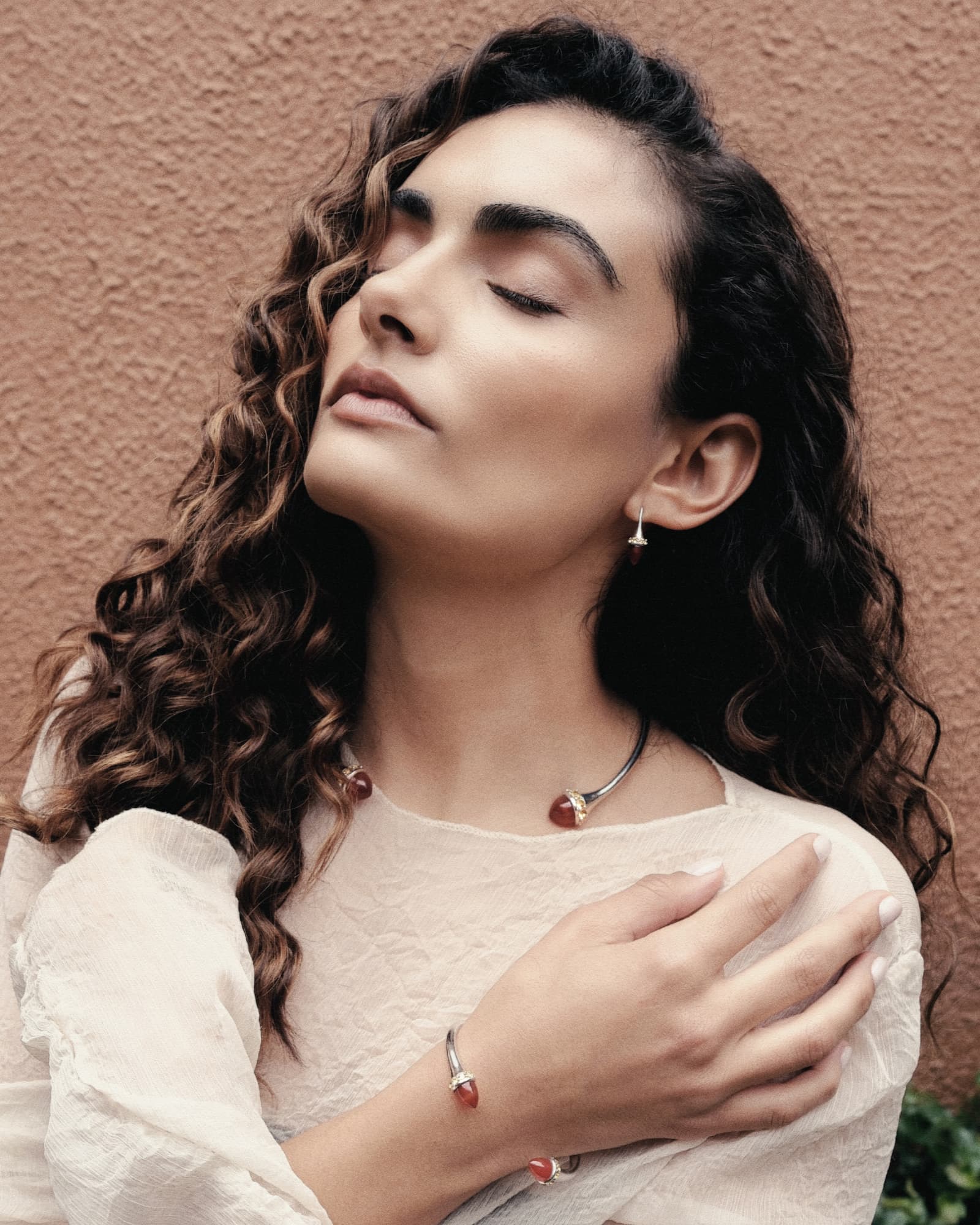 Woman with long, wavy hair wearing a white top against a beige wall.
