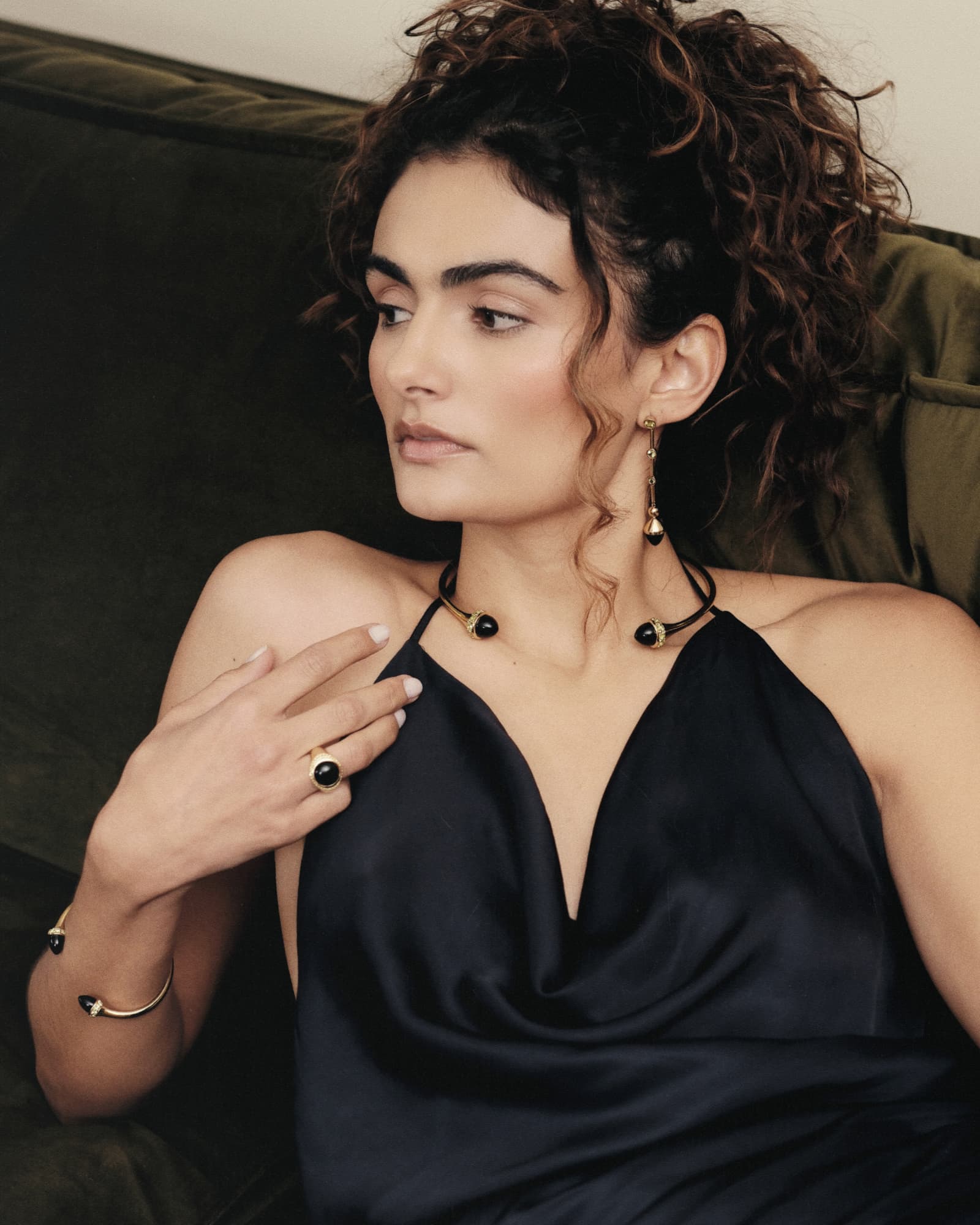 Woman wearing a black dress and jewelry, sitting on a green couch.
