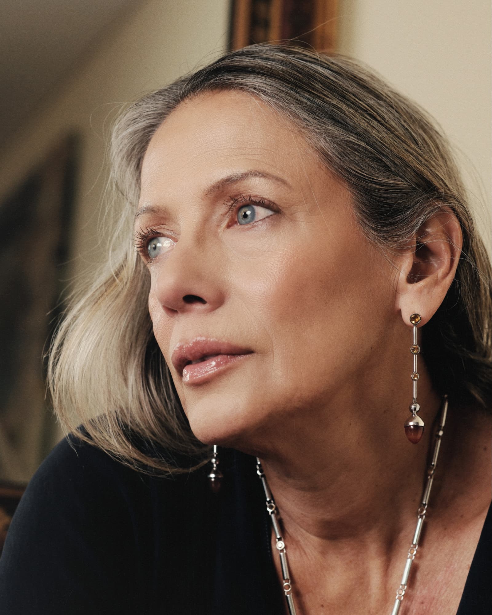 Close-up of a woman wearing earrings and a necklace, with a blurred indoor background.